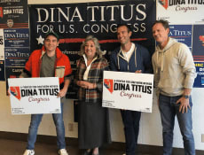 Jon Lovett, Rep. Dina Titus, Jon Favreau, and Tommy Vietor posing for a photo in front of a wall of Dina Titus for Congress posters.