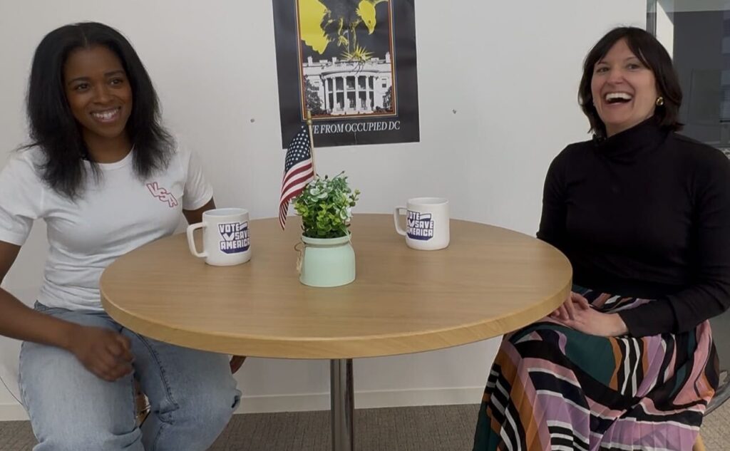 Shaniqua McClendon and Hannah Fried smile off camera while seated at a wooden table.