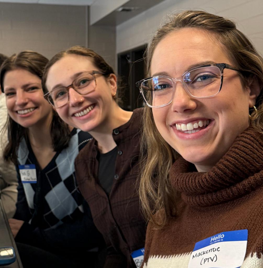 A woman smiles while taking a selfie next to two other women. She has a name tag that reads Hello my name is Mackenzie (PTV).
