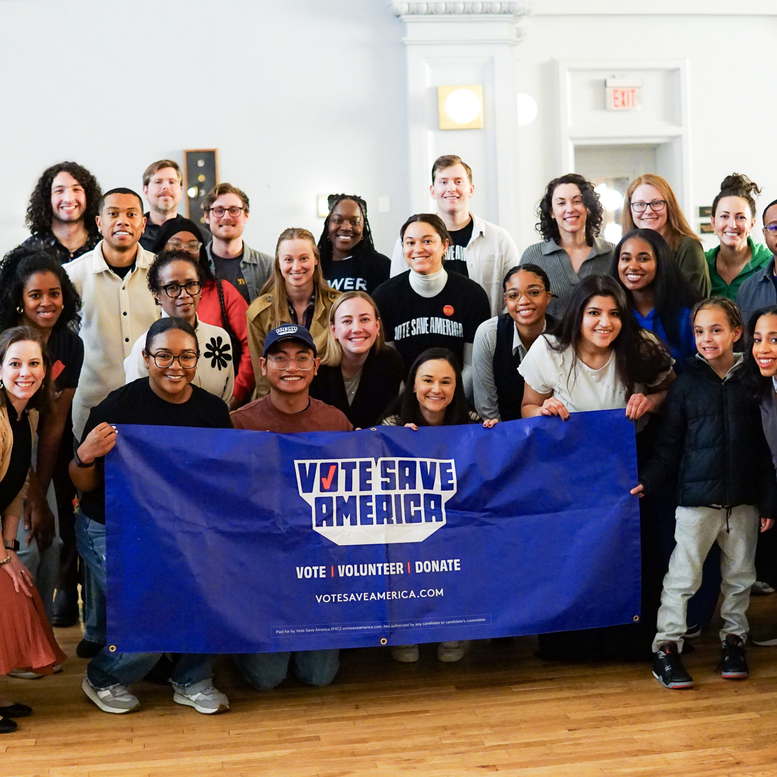 A group of volunteers of different skin tones smile to camera holding a blue banner that reads 'Vote Save America.'
